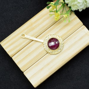 Puede incluir: Una pinza para el pelo dorada con un diseño redondo y adornado que presenta una piedra preciosa de color rojo intenso en el centro.