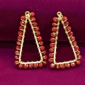May include: A pair of gold triangle earrings with red beads. The earrings are made with wire and have a geometric design.