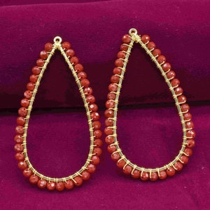 Pode incluir: Um par de brincos dourados com contas vermelhas. Os brincos têm a forma de uma lágrima e são feitos com uma base de arame.