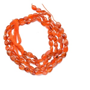May include: Strands of oval-shaped, polished carnelian beads in shades of orange and red. The beads are strung together with orange thread, creating a natural and earthy aesthetic. Ideal for jewelry making.