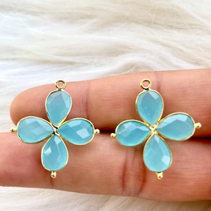 Könnte beinhalten: Zwei goldfarbene blütenförmige Anhänger mit hellblauen Edelstein-Akzenten. Jeder Anhänger hat vier Blütenblätter und eine Schlaufe zum Anbringen an Schmuck.
