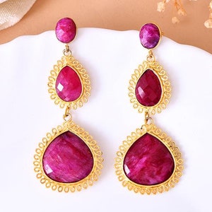 May include: A pair of gold earrings with pink gemstone drops. Each earring features three pink gemstones, two of which are surrounded by a delicate gold filigree design.