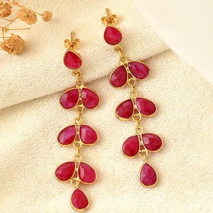May include: A pair of gold-colored drop earrings featuring vibrant red gemstones. Each earring has a series of three petal-shaped stones, with a single teardrop-shaped stone at the bottom. The earrings are displayed on a white, textured surface.