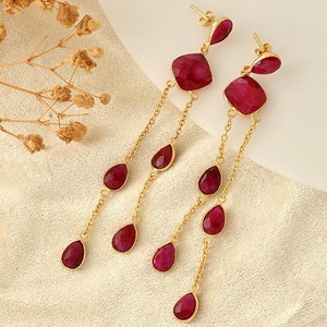 Könnte beinhalten: Ein Paar lange, baumelnde Ohrringe mit roten Edelstein-Tropfen und einem größeren quadratischen Edelstein oben. Die Ohrringe sind aus goldfarbenem Metall.