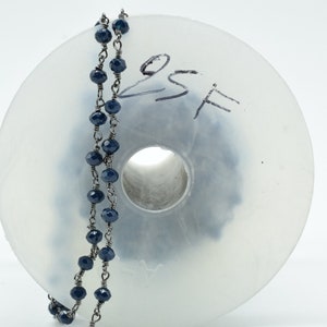 Puede incluir: Un carrete de cuentas facetadas de color azul oscuro enhebradas en una cadena de plata. El carrete es de pl&aacute;stico transparente y tiene el texto escrito a mano "8SF".