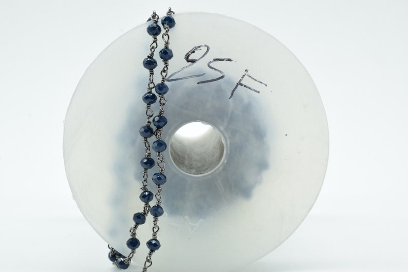 Puede incluir: Un carrete de cuentas facetadas de color azul oscuro enhebradas en una cadena de plata. El carrete es de pl&aacute;stico transparente y tiene el texto escrito a mano "8SF".