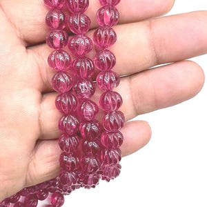 Op de afbeelding: Een streng gepolijste, doorschijnende, roze edelsteen kralen. De kralen hebben een unieke, gesneden pompoenvorm. De kralen zijn aan elkaar geregen, waardoor een ketting of armband ontstaat. De achtergrond is wit.