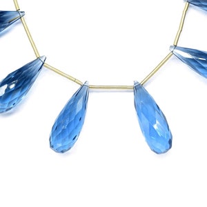 May include: A strand of blue topaz gemstone beads, each bead is faceted and shaped like a teardrop. The beads are strung on a gold wire.