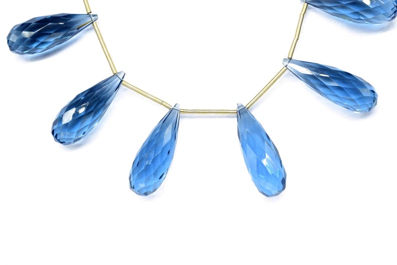 May include: A strand of blue topaz gemstone beads, each bead is faceted and shaped like a teardrop. The beads are strung on a gold wire.