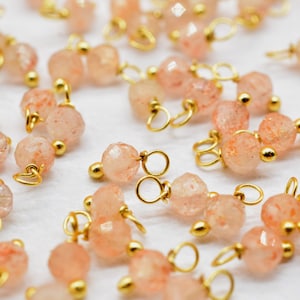 Könnte beinhalten: Eine Sammlung von goldfarbenen Metallperlenkappen mit daran befestigten Sunstone-Perlen. Die Perlen sind hellpfirsichfarben mit einem schimmernden, durchscheinenden Finish.