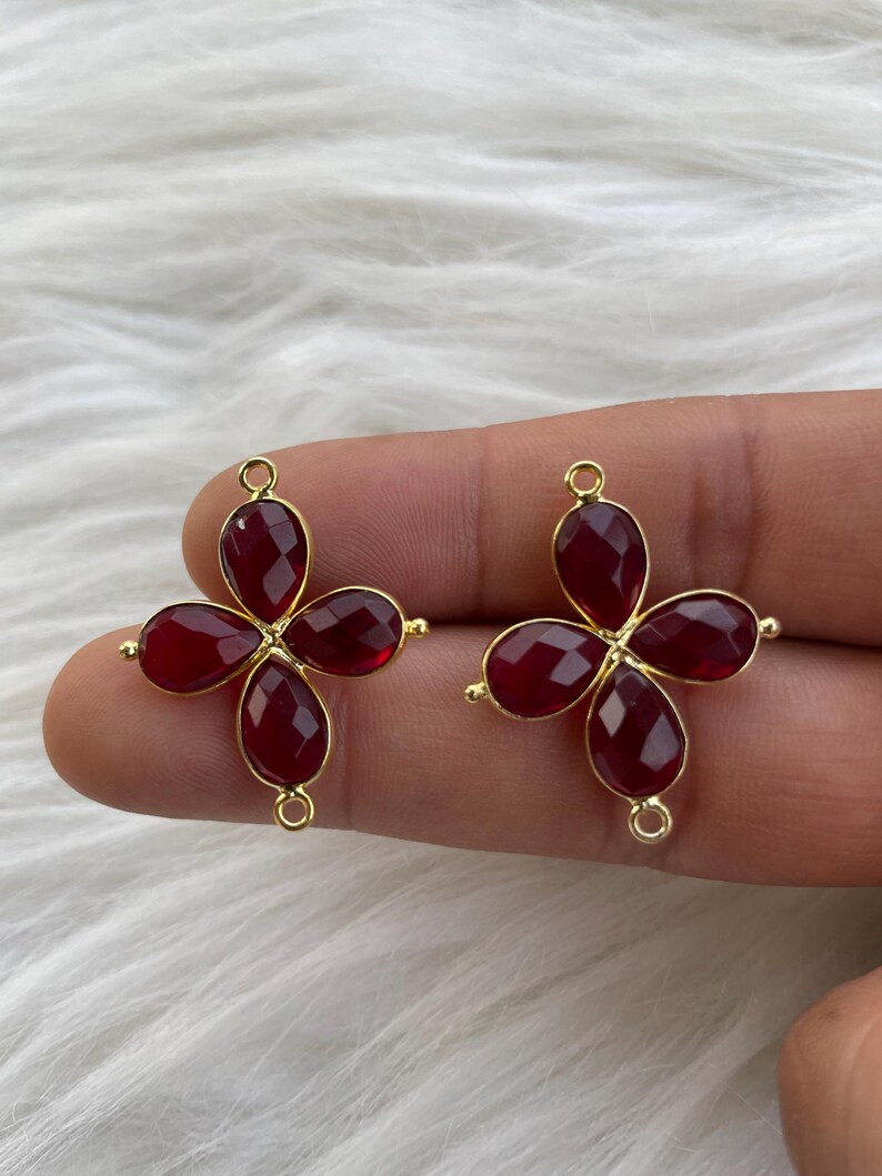 May include: Two gold-tone metal flower-shaped charms with red gemstone accents. Each charm has four red gemstone petals and a loop for attaching to a necklace or bracelet.