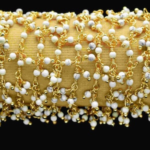 May include: A close-up of a gold chain with white beads. The chain is made of small, delicate links and the beads are round and smooth. The chain is lying on a brown surface.