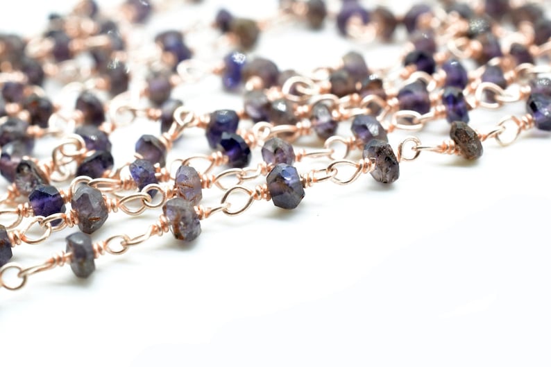 May include: A close-up of a chain made of small, faceted, purple gemstones and rose gold wire. The chain is on a white background.