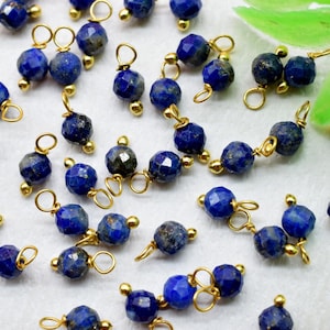 May include: A collection of faceted, deep blue lapis lazuli beads with gold-colored metal accents. Each bead is connected with a small loop, suggesting they are components for jewelry making. The beads are scattered on a light surface.