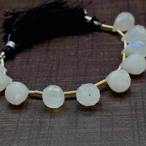 Puede incluir: Una hebra de cuentas de piedra lunar blanca con un alambre dorado enhebrado. Las cuentas son facetadas y tienen forma de pera. La hebra est&aacute; terminada con un borla negra.