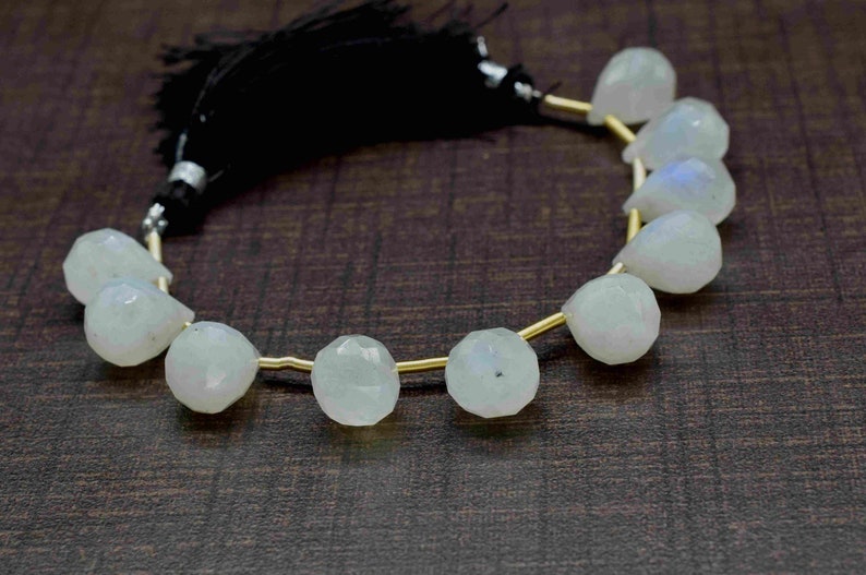 Puede incluir: Una hebra de cuentas de piedra lunar blanca con un alambre dorado enhebrado. Las cuentas son facetadas y tienen forma de pera. La hebra est&aacute; terminada con un borla negra.