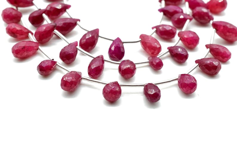 May include: A strand of ruby red gemstone beads in a teardrop shape. The beads are strung on a silver wire.