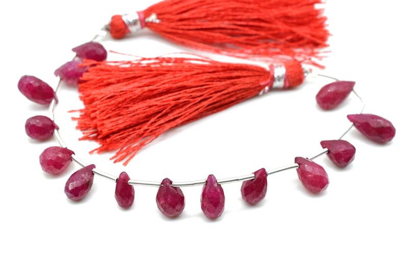May include: A strand of ruby gemstone beads in a teardrop shape, strung on a silver wire. The beads are a deep red color and are arranged in a circular pattern. Two red tassels are included.