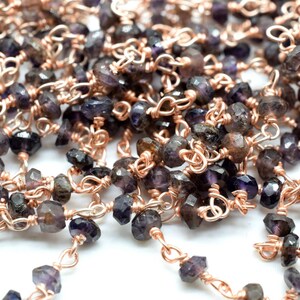 May include: A close-up of a chain made of small, faceted, dark purple gemstones and rose gold wire. The chain is made of individual links, each with a gemstone and a small rose gold loop.