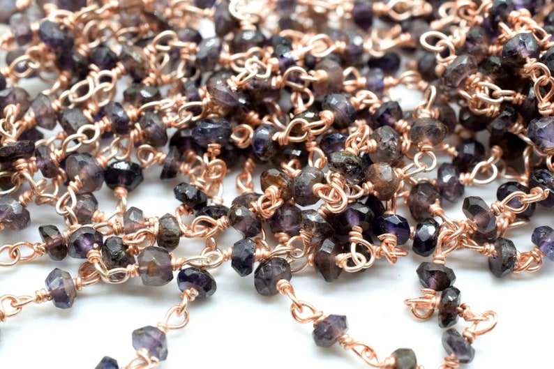 May include: A close-up of a chain made of small, faceted, dark purple gemstones and rose gold wire. The chain is made of individual links, each with a gemstone and a small rose gold loop.