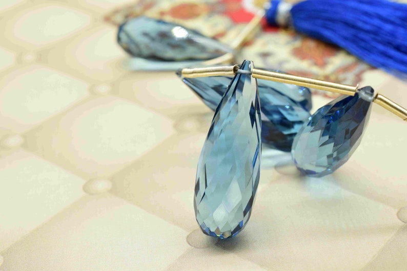 May include: Three teardrop-shaped blue topaz gemstones with gold-toned jump rings. The gemstones are faceted and have a light blue color.