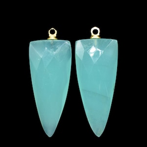Puede incluir: Dos colgantes de piedra preciosa azul claro con anillas de salto de oro. Los colgantes son facetados y tienen una punta puntiaguda.