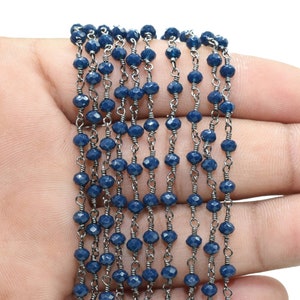 Puede incluir: Una cadena de cuentas facetadas de color azul oscuro con eslabones de cadena plateados. La cadena est&aacute; dispuesta en l&iacute;nea recta sobre una mano.