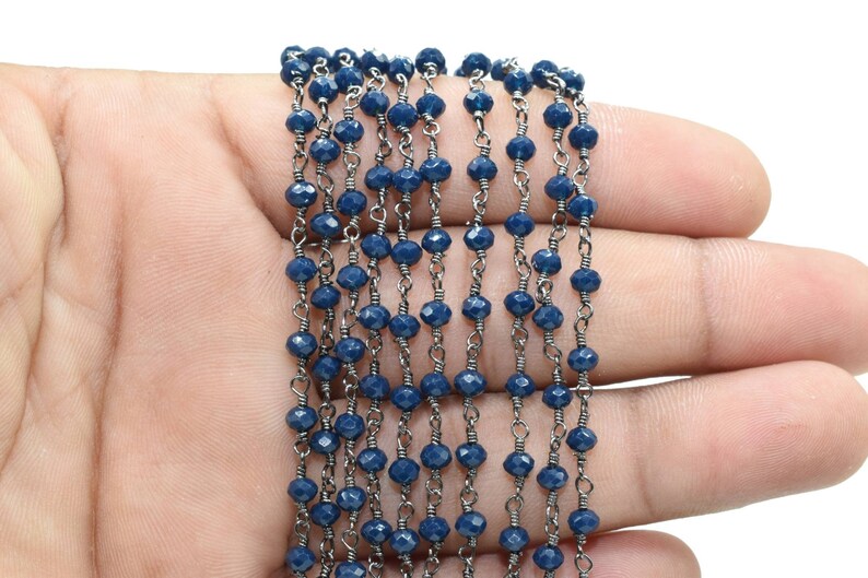 Puede incluir: Una cadena de cuentas facetadas de color azul oscuro con eslabones de cadena plateados. La cadena est&aacute; dispuesta en l&iacute;nea recta sobre una mano.
