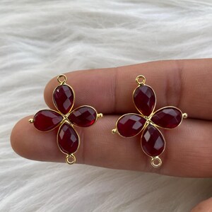 May include: Two gold-tone metal flower-shaped charms with red gemstone accents. Each charm has four red gemstone petals and a loop for attaching to a necklace or bracelet.