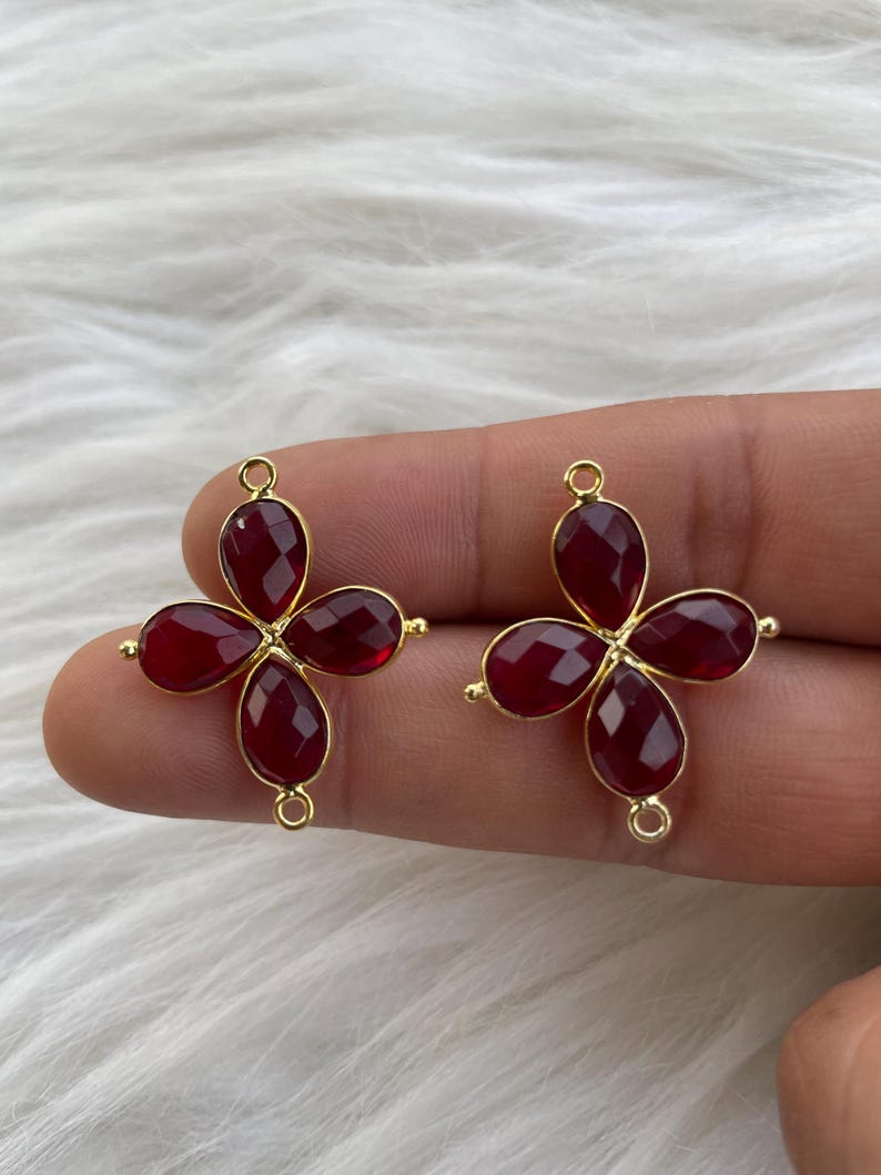 May include: Two gold-tone metal flower-shaped charms with red gemstone accents. Each charm has four red gemstone petals and a loop for attaching to a necklace or bracelet.