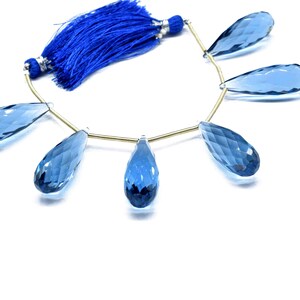 May include: A strand of blue topaz gemstone beads, each bead is faceted and shaped like a teardrop. The beads are strung on a gold wire and have a blue tassel at the end.