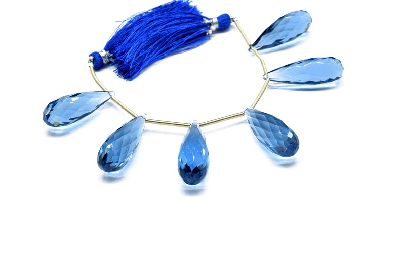 May include: A strand of blue topaz gemstone beads, each bead is faceted and shaped like a teardrop. The beads are strung on a gold wire and have a blue tassel at the end.