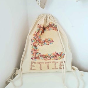 May include: A beige drawstring bag with a floral design and the name "ETTIE" printed on the front.
