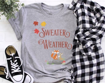 May include: A gray t-shirt with the words "Sweater Weather" in a decorative font, accompanied by illustrations of autumn leaves and a fox with a rabbit. A black and white plaid shirt and black sneakers are also in the image.