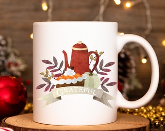 May include: White ceramic mug with a fall-themed design. The design includes a red teapot, a slice of pie, a light green pumpkin, and the word "Grateful" on a banner. The mug is on a wooden surface.