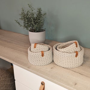 Puede incluir: Conjunto de tres cestas de almacenamiento de crochet beige con etiquetas de cuero. Las cestas están apiladas sobre una superficie de madera clara.