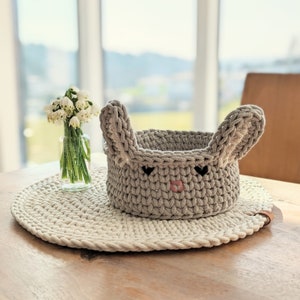 May include: A handmade gray crochet bunny basket with upright ears, black eyes, and a pink nose. The basket sits on a round, cream-colored placemat. A small glass vase with white flowers is to the left of the basket.
