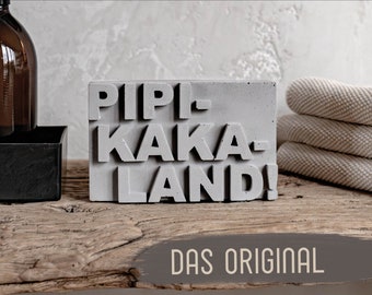 May include: A gray concrete block with the words "PIPI-KAKA-LAND!" in raised letters. The block is sitting on a wooden surface with the words "DAS ORIGINAL" in white letters on a brown background.