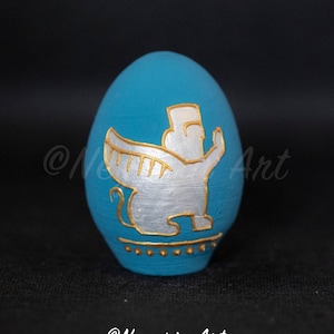 May include: A turquoise egg-shaped decorative item with a silver and gold design. The design features a stylized figure with wings. The base of the egg has a gold border. The artist's name, "@Negarin Art", is written on the egg.