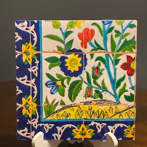 May include: A ceramic tile with a floral design. The tile features blue, yellow, green, and red flowers on a white background. The tile is framed with a blue border.