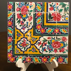 May include: A square tile with a colorful floral pattern. The tile is decorated with blue, green, yellow, and red flowers on a white background. The tile is framed by a border of blue, green, and yellow.