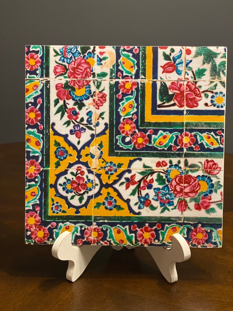 May include: A square tile with a colorful floral pattern. The tile is decorated with blue, green, yellow, and red flowers on a white background. The tile is framed by a border of blue, green, and yellow.