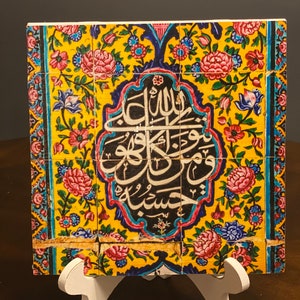 May include: A colorful ceramic tile with intricate floral patterns and Arabic calligraphy in black. The tile is mounted on a white easel.