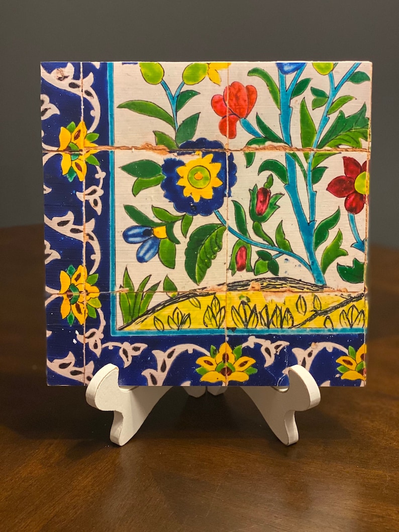 May include: A ceramic tile with a floral design. The tile features blue, yellow, green, and red flowers on a white background. The tile is framed with a blue border.