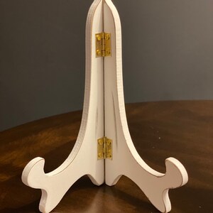 May include: White wooden easel stand with gold hinges. The stand is folded and is sitting on a wooden surface.