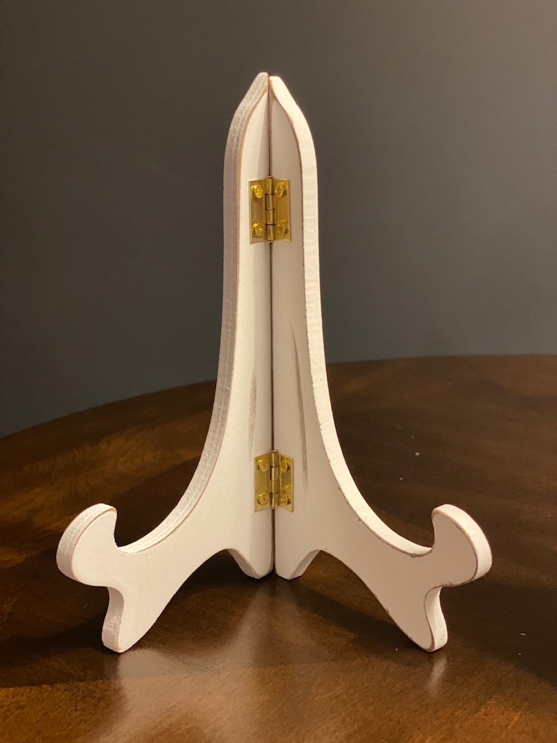 May include: White wooden easel stand with gold hinges. The stand is folded and is sitting on a wooden surface.