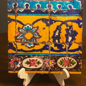 May include: A square tile with a colorful mosaic design. The tile features blue, yellow, pink, and black floral patterns. The tile is displayed on a white easel.