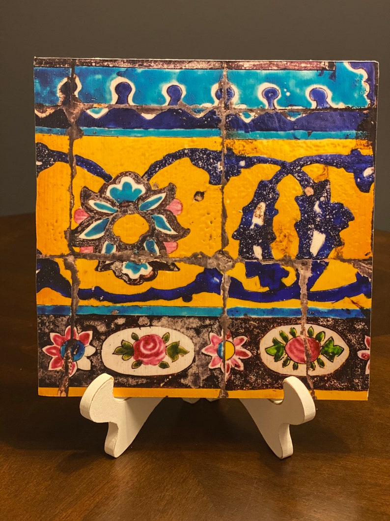 May include: A square tile with a colorful mosaic design. The tile features blue, yellow, pink, and black floral patterns. The tile is displayed on a white easel.
