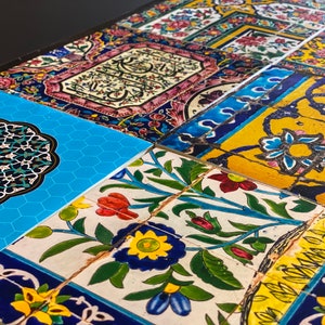 May include: A close-up of a table top covered in colorful, patterned tiles. The tiles feature floral designs, geometric shapes, and Arabic script. The tiles are arranged in a patchwork pattern.
