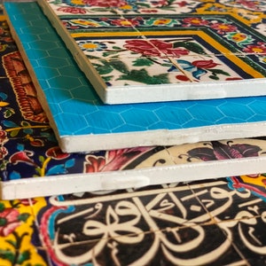 May include: Close-up of the edges of three ceramic tiles with intricate floral patterns and Arabic calligraphy. The tiles are stacked on top of each other, with the top tile featuring a blue and white geometric pattern.